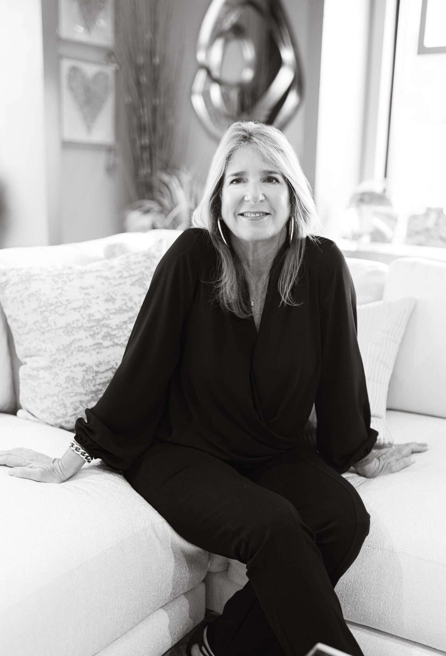Modern woman sitting on a white sofa in an art-filled living room, smiling, representing interior design and home comfort.