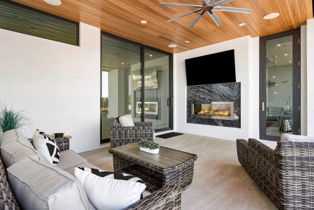 Covered outdoor living room with wood ceiling, woven seating, wall-mounted TV, and built-in fireplace.