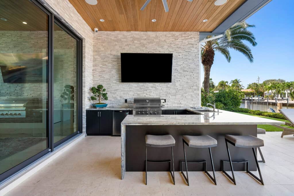 Luxury covered outdoor kitchen with built-in grill, bar seating, stone accent wall, wood ceiling, and waterfront views.